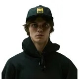 Gorra snapback negra con visera plana y parche frontal rectangular de color dorado con un diseño gráfico abstracto que simula dos caras o formas simétricas.