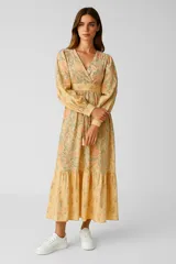 Vestido largo de algodón color amarillo con estampado floral en tonos naranja y verde. Tiene escote en V cruzado, mangas largas abullonadas con puños elásticos, cintura ajustada con lazo y falda con volantes.