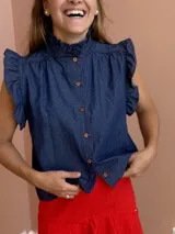 Blusa de denim azul con cuello alto con volados, mangas cortas con volados y cierre frontal con botones.