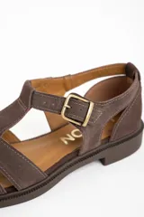 Sandalia cangrejera de gamuza color marrón oscuro, con puntera cerrada y tiras entrecruzadas. Posee tira de ajuste con hebilla metálica dorada en el tobillo y suela de goma plana.