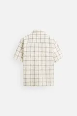 Camisa de manga corta color crudo con estampado de cuadros en tonos grises y marrones. Tiene cuello bowling, corte relaxed fit, aberturas laterales en el bajo y cierre frontal con botones.
