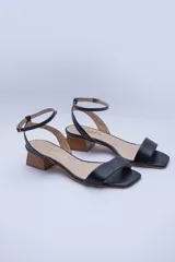 Sandalias de cuero negro con tira delgada sobre los dedos, pulsera al tobillo con hebilla dorada y taco geométrico color madera.