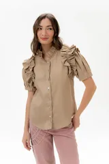 Camisa de lino color beige, de manga corta con volados amplios y cuello alto con volados. Presenta un frente abotonado.