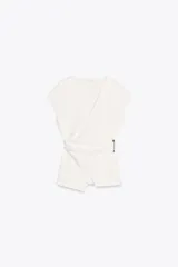 Top blanco de escote pico cruzado y manga sisa caída. Tejido con textura y broche metálico lateral. Bajo asimétrico.