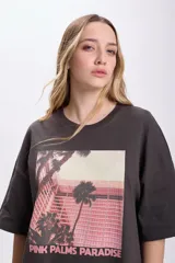 Remera manga corta oversize color gris oscuro con estampa fotográfica de palmeras y edificio rosado.