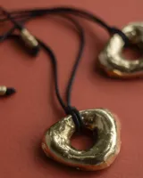 Collar con dije dorado irregular con agujero central, colgando de cordón negro ajustable.