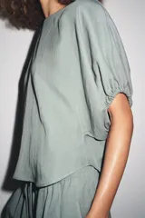 Blusa de cuello redondo color verde, confeccionada en hilatura con mezcla de lyocell. Manga corta abullonada ajustable con lazadas. Bajo redondeado.