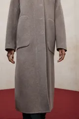 Abrigo largo de lana reciclada en color negro, con diseño minimalista, cuello redondo y cierre frontal con botones ocultos. Presenta aberturas laterales, bolsillos frontales tipo parche y mangas largas.