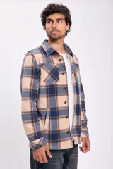 Sobrecamisa de tejido grueso a cuadros, con cuello camisero, botones frontales y bolsillo de parche en el pecho. El diseño combina tonos beige tostado, azul marino y marrón.