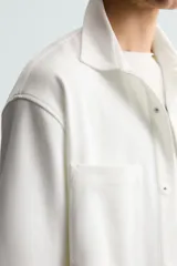 Sobrecamisa negra de tejido con elasticidad, cuello solapa y manga corta. Tiene dos bolsillos de plastrón en el pecho y cierre frontal con botones a presión.