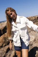 Chaqueta de denim liviana en color beige con detalle de volados en el frente, cuello camisero y bolsillos funcionales. Presenta un calce relajado y cierre frontal mediante botones.