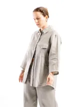 Camisa de lino color gris claro, con mangas 3/4, cuello clásico, cierre frontal con botones, bolsillo en el pecho izquierdo y canesú con pliegue en la espalda. Corte holgado.