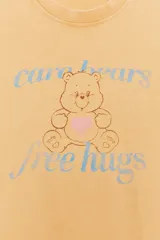Camiseta color amarillo con efecto lavado, cuello redondo y mangas cortas. Estampado frontal con un oso de los Care Bears™ y la frase "free hugs".