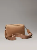 Bandolera Calvin Klein color tostado, con correa ancha de tela con logo de la marca y correa fina de cuero sintético.