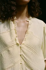 Blusa color crema de escote pico con volantes y manga larga. Detalle de jaretas y entredoses a tono. Cierre frontal con botones forrados a tono.