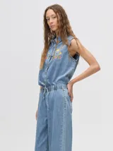 Mono largo de jean celeste con corte holgado, cintura elastizada con cordón ajustable, sin mangas, cuello camisero y cierre frontal con botones. Presenta bordados de motivos playeros en el pecho.