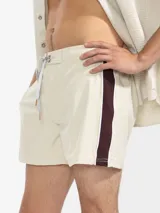 Short de baño color beige con una franja vertical marrón en el lateral. Tiene cintura elástica con cordón ajustable, bolsillos laterales y trasero con cierre, y suspensor de malla fina hipoalergénica.