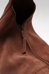 Bolso tipo hobo de gamuza negra, con forma holgada y cierre principal de cremallera. Se lleva al hombro con dos asas de correa delgadas sujetas con argollas metálicas plateadas.