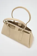 Bolso tipo tote color beige, con doble asa corta y cierre superior con botón imantado. Incluye un compartimento interno con cierre de cremallera.