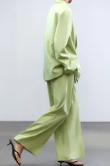 Pantalón verde claro de tiro alto con trabillas y botón en la cintura. Tiene pliegues, bolsillos laterales y falsos bolsillos de vivo en la espalda. El cierre frontal es con cremallera, botón interior y ganchos metálicos.