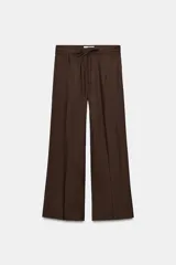 Pantalón marrón de tiro alto con bolsillos laterales ocultos en costuras, pliegues en delantero y cinturón doble en mismo tejido. Cierre frontal con cremallera y gancho metálico.