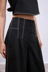 Pantalón negro de tiro alto, corte ancho y largo, con pliegues frontales y pespuntes contrastantes en blanco en la cintura y bolsillos.