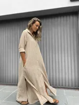 Vestido camisero largo con estampado de rayas verticales en beige y blanco. Presenta cuello camisero, cierre frontal con botones, mangas largas y tajos laterales en el ruedo.