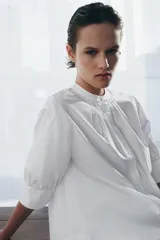 Camisa blanca de algodón con cuello redondo y manga corta abullonada. Presenta un canesú con pliegues verticales en la parte delantera y cierre frontal con botones ocultos. El bajo es redondeado.