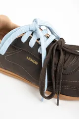 Zapatilla de cuero marrón oscuro, estilo deportivo retro, con suela de goma color caramelo. Presenta costuras decorativas en el cuerpo y el nombre "LEMON" grabado en dorado en el lateral. Se muestra con dos cordones anudados: uno celeste claro y otro marrón oscuro.