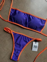 Bikini de lycra violeta oscuro con ribetes naranja. El corpiño tiene doble tela y aberturas para copas. Los breteles y tiras de la bombacha son regulables. La bombacha es colaless.