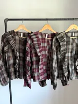 Camisa de tela escocesa liviana, color gris oscuro con líneas en blanco, azul marino y salmón. Presenta un diseño con volados verticales en el frente y mangas abullonadas.