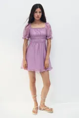 Vestido corto lila con escote cuadrado, mangas cortas abullonadas y fruncido elástico bajo el busto. Presenta volados en el escote, mangas y ruedo.