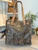 Cartera tipo tote de cuero color marrón con efecto craquelado, con flecos en la parte frontal y cierre con broche metálico. Tiene doble asa de hombro.