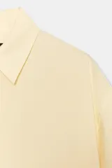 Camisa color amarillo pálido de cuello solapa y manga corta con dobladillo. Tiene bajo asimétrico con aberturas laterales y cierre frontal con botones ocultos.