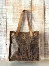 Cartera tipo tote de cuero color marrón con efecto craquelado, con flecos en la parte frontal y cierre con broche metálico. Tiene doble asa de hombro.