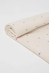 Pashmina color beige con pequeños puntos bordados en varios colores (rojo, azul, amarillo, verde, violeta). Tiene flecos en los extremos y un parche de cuero sintético marrón con un logo grabado.
