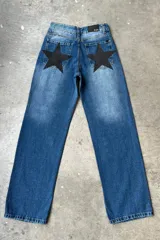 Jean de mezclilla azul medio con efecto desgastado y apliques de estrellas negras de tela texturizada en los bolsillos traseros. Presenta etiqueta de marca negra con texto blanco en la cintura trasera.