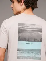 Remera color beige de algodón regenerativo con cuello redondo, mangas cortas y logo CK bordado en el pecho. Presenta un diseño de paisaje gráfico en el frente.
