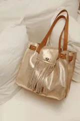 Bolso tipo tote de cuero dorado con efecto metalizado y flecos. Tiene doble asa de cuero marrón y bolsillo frontal con cierre de broche.