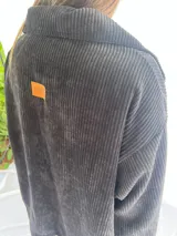 Buzo de pana fina color gris oscuro, con cuello camisero y corte holgado. Presenta un parche de cuero naranja con el logo 'Miza' en la espalda.