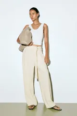 Pantalón sastrero color crudo, de tiro alto con elástico oculto en la cintura y cierre decorativo. Cinturón cosido con anillas en D de metal en un lateral. Piernas ajustadas con pinzas para estilizar la silueta.