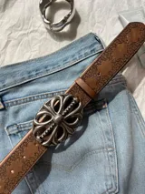 Cinturón marrón de cuero con estampado y hebilla plateada con diseño floral. Incluye un segundo cinturón de cadena plateada con dijes circulares.