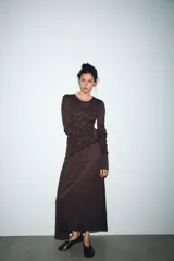 Vestido midi color marrón oscuro, con cuello redondo y manga larga. Presenta frunces diagonales en la parte superior delantera y en las mangas, y un bajo asimétrico.