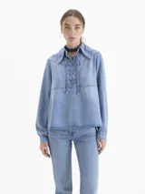 Camisa de denim azul claro, manga larga, con cuello clásico y escote ajustable mediante cordón que pasa por ojalillos metálicos.