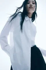 Camisa blanca oversized de popelina de algodón, con cuello clásico, botones frontales, hombros caídos, mangas largas con puños abotonados, canesú con pliegue en la espalda y bajo redondeado.