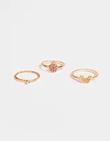 Set de tres anillos dorados con strass: uno con diseño de flor rosa, otro con diseño de mariposa y otro con una pequeña piedra verde.