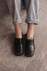 Mocasín de cuero negro con cordones y tachas metálicas en la puntera.