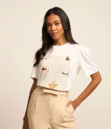 Remera blanca de corte slim con cuello redondo y mangas cortas abullonadas. Presenta un estampado frontal con diseño de picnic en tonos beige, con detalles bordados de abeja, cesta, hongo y texto en francés.