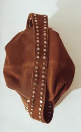 Bolso de hombro tipo hobo, color camel, confeccionado en gamuza o material similar con acabado afelpado. Presenta detalles de tachas metálicas plateadas a lo largo de los bordes laterales y en la correa de hombro.