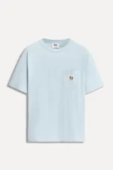 Camiseta celeste de algodón con cuello redondo y manga corta. Tiene un bolsillo de parche en el pecho con el logo del 50 aniversario de la marca.